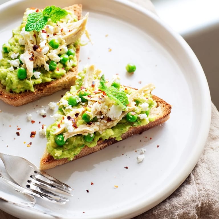 Close-up of vibrant green pea mash and tender chicken on crisp toast, sprinkled with feta and mint.